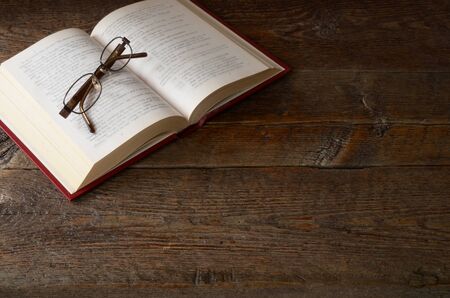 A top view image of a single open book and pair of old eye glasses on a old wooden table top.の写真素材