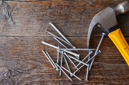 A top view image of an old used yellow hammer and a pile of nails on a wooden workbench.