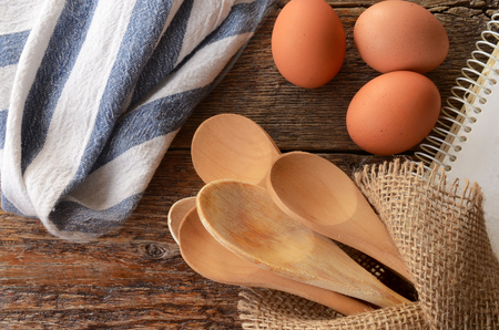 A top view image of wooden mixing spoons, brown eggs, and an old cookbook on a kitchen table.の写真素材