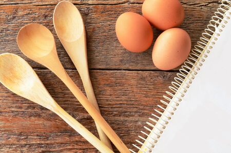 A top view image of wooden mixing spoons, brown eggs, and an old cookbook on a kitchen table.の写真素材