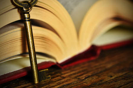 A close up image of an open book with antique key on a dork wooden desk top.