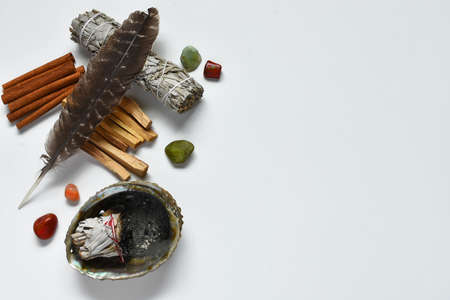 A top view image of white sage, palo santo, healing crystals, and a sacred feather on a white...