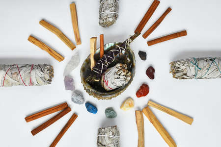 A top view image of white sage, palo santo, healing crystals, and a sacred feather on a white...