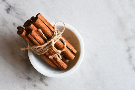 A top view image of several cinnamon sticks in a white porcelain bowl on a marble table top.