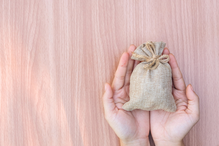 Hand holding money sack or bag with coins on wooden background. Save and investment money for prepare in the future - concept of saving money.の写真素材