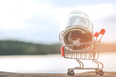 Wooden house in a small supermarket trolley and coins Inside a glass jar, credit, tax - financial conceptの写真素材