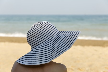 white tanned woman head closeup in stripped broad beach hat with sea beach background, back viewの写真素材