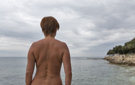 attractive tanned naked middle aged woman standing back to the camera on the background of the overcast Mediterranean sea and the coastの写真素材