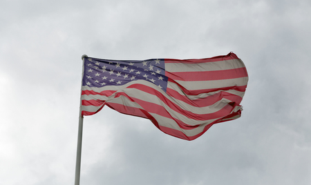 Faded and tattered American flag waving against a cloudy skyの写真素材