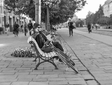 KOSICE, SLOVAKIA - OCTOBER 02, 2017: Beautiful young woman sits with smartphone and bicycle on the bench in Old Town in black and white. Kosice city was the European Capital of Culture in 2013.のeditorial素材