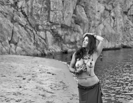 young caucasian beautiful naked Amazon woman on the sand river beach in front of rocks. black and...