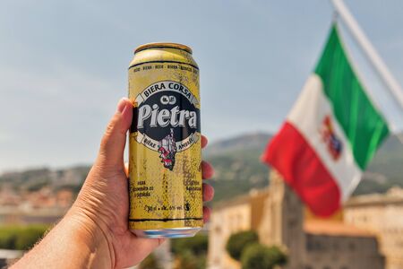 BASTIA, CORSICA, FRANCE - JULY 23, 2019: White man's hand holds a can of Pietra Corsican beer closeup with Italian flag and Bastia cityscape in the background.のeditorial素材