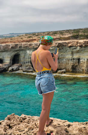 Ayia Napa, Cyprus - May 27, 2021: Young beautiful woman use smartphones in Cape Greco peninsula park. It is mountainous peninsula with a national park, rock paths and turquoise lagoon.のeditorial素材