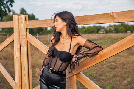 Beautiful young brunette caucasian woman wearing black leather cloth standing at the ranch gate.の写真素材