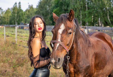 Beautiful young brunette caucasian woman in black leather cloth standing near the horse, closeup.の写真素材