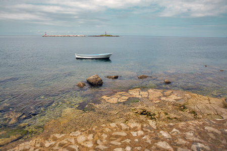 Small white boat floating on calm sea near rocky shore. Lighthouse on Barbaran island in the background. Peaceful coastal scene, clear water, summer travel and vacation concept. Porec, Istria, Croatiaの写真素材