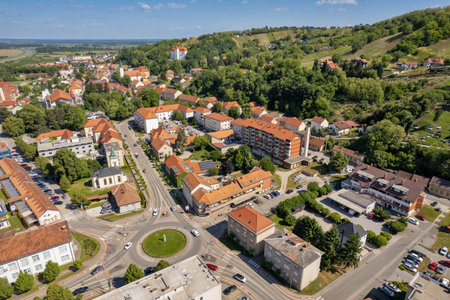 Drone aerial photograph of picturesque Slovenian town Lendava featuring traditional red clay tile rooftops, central traffic roundabout, residential buildings surrounded by lush green hills and forest.の写真素材