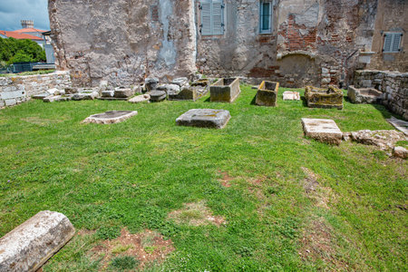 Ancient Roman archaeological site in Porec, Croatia showing excavated stone foundations, scattered ruins and artifacts on green grass with medieval buildings in background.の写真素材