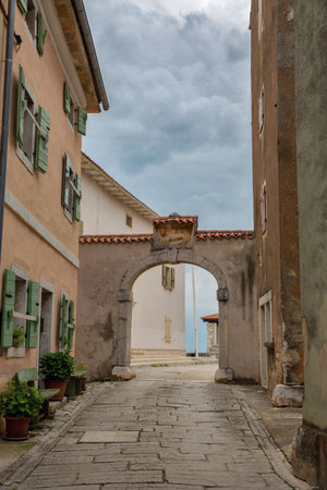 Ancient archway with decorative carved relief connects traditional Mediterranean buildings with green shutters along cobblestone street. Historic Istrian village Visnjan, Croatia.の写真素材