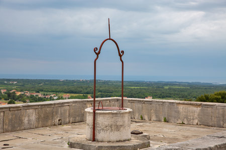 Ancient water stone well with decorative red iron mechanism on medieval fortress terrace overlooking green Istrian countryside and scattered villages below. Visnjan, Croatia.の写真素材