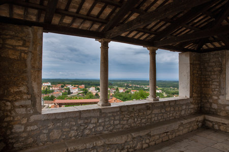 Scenic view of Visnjan, Croatia, framed by stone pillars and arches of an old balcony, with tiled rooftops and green landscape under a cloudy sky.の写真素材
