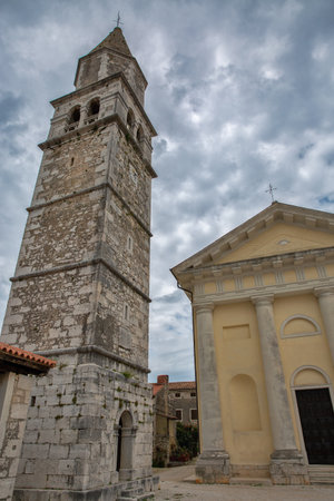 Medieval stone bell tower stands beside neoclassical Church of the Sts. Cyricus and Julitta with columned portico under cloudy sky in Croatian Istrian village Visnjan.の写真素材