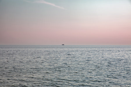 Peaceful seascape with lone ship on tranquil waters during golden hour. Pink blue evening sky.