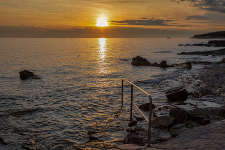 Stunning golden hour seascape featuring rocky shore with metal swimming ladder leading into calm waters. Sun creates beautiful reflection path across the sea.の写真素材