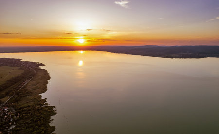 Breathtaking sunset over Lake Balaton with vibrant orange and yellow sky reflecting on calm water surface. Perfect golden hour lighting creates stunning Hungarian lakeside landscape scene.の写真素材