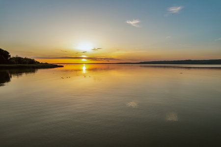 Breathtaking sunset over Lake Balaton with vibrant orange and yellow sky reflecting on calm water surface. Perfect golden hour lighting creates stunning Hungarian lakeside landscape scene.の写真素材