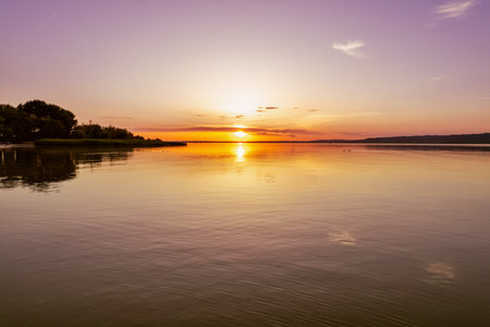 Breathtaking sunset over Lake Balaton with vibrant orange and yellow sky reflecting on calm water surface. Perfect golden hour lighting creates stunning Hungarian lakeside landscape scene.の写真素材