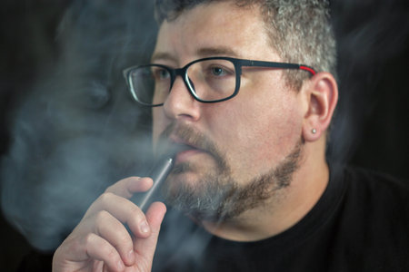 Studio portrait of young Caucasian man smoking an electronic cigarette and wearing glasses and...