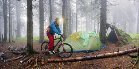 Ukraine, Vorohta- June 3, 2016: rain mist rider on a mountain bike prepares for spending the night in a tent near the old hut of moss in the wild alpine spruce forest among the windbreaksのeditorial素材