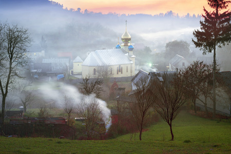 Carpathian valleys with smoke Christian church in the Carpathian Mountains, Ukraine - villagers harness fallen leaves and stoke the furnace, the absence of wind delays the mist over the villageの写真素材