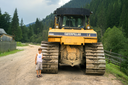 Ukraine, Verkhovyna, 15 July 2015:  boy from the Carpathian village is next  large iron Caterpillar tractor paving the way to their village of construction of hydroelectric power station damのeditorial素材