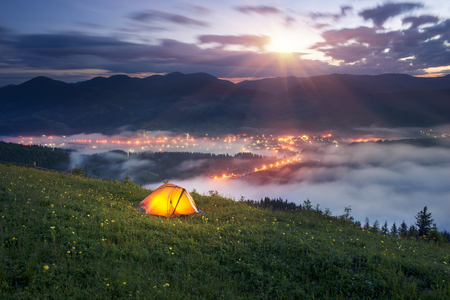 tent is illuminated at the top of the mountain, the waves of the misty sea float in the valley against the backdrop of high peaks covered in forests. Below is a mountain village shiningの写真素材