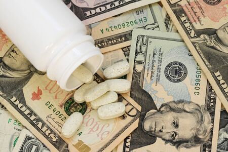 white pill bottle spilling drug pills tablets out onto a background of US paper money illustrating the high cost of drugsの写真素材