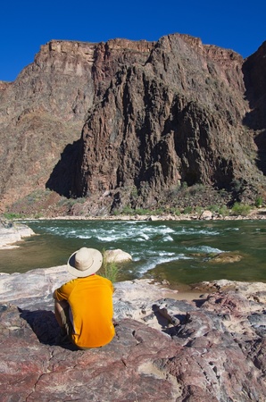back view of a man sitting on a rock on the edge of the Colorado River at the bottom of the Grand Canyonの写真素材