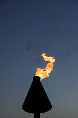 metal tiki torch with yellow flame before a blue skyの写真素材