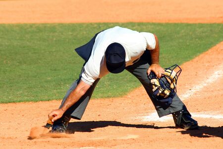 Baseball umpire dusting off dirt from home plate with a brush with his mask in his other handの写真素材