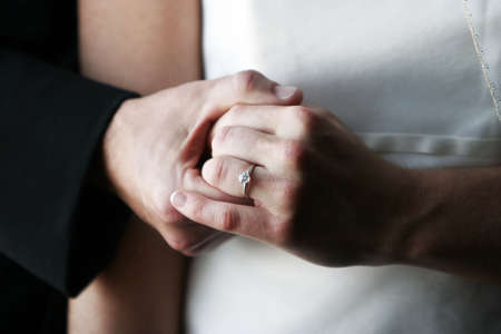 Groom holding the left hand of his bride displaying her engagement ring, white wedding gress and black suitの写真素材