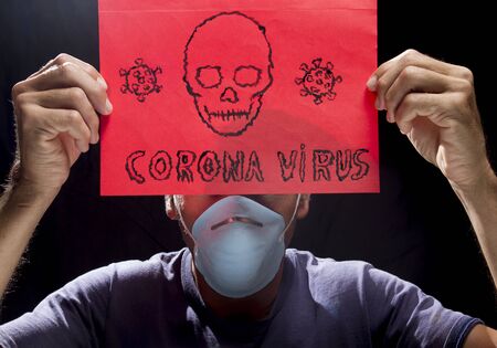 man wearing blue surgery mask holding red sign of coronavirus and skeleton in a black background