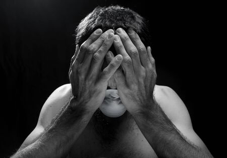 crestfallen or worried white man with hands in head and surgical mask in trouble in a dark room or black background.concept of sadnessの写真素材