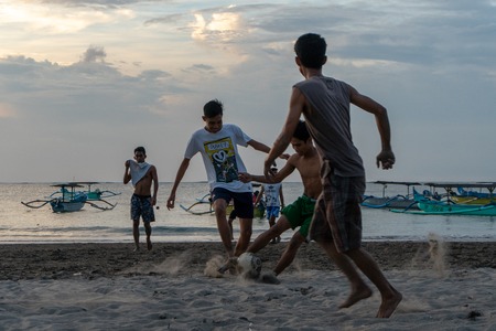 BADUNG,BALI/INDONESIA-APRIL 02 2019: Asian Teenager play football or soccer at Kuta beach with sunset or golden hours background.The sky is cloudy and some traditional boat which is called jukung, anchored thereのeditorial素材