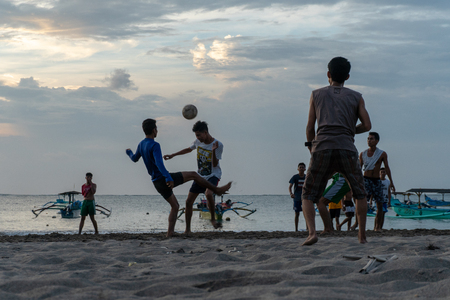 BADUNG,BALI/INDONESIA-APRIL 02 2019: Asian Teenager play football or soccer at Kuta beach with sunset or golden hours background.The sky is cloudy and some traditional boat which is called jukung, anchored thereのeditorial素材