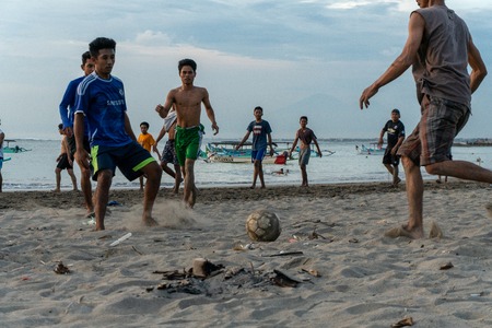 BADUNG,BALI/INDONESIA-APRIL 02 2019: Asian Teenager play football or soccer at Kuta beach with sunset or golden hours background.The sky is cloudy and some traditional boat which is called jukung, anchored thereのeditorial素材