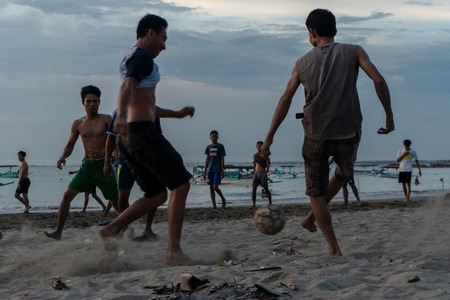 BADUNG,BALI/INDONESIA-APRIL 02 2019: Asian Teenager play football or soccer at Kuta beach with sunset or golden hours background.The sky is cloudy and some traditional boat which is called jukung, anchored thereのeditorial素材