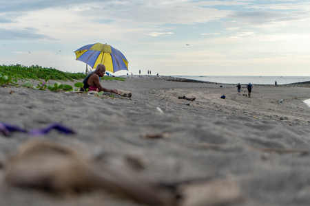 BADUNG,BALI/INDONESIA-APRIL 02 2019: Elderly man sits on the sand and enjoy sunbathing at Kuta beach with blue yellow umbrellaのeditorial素材