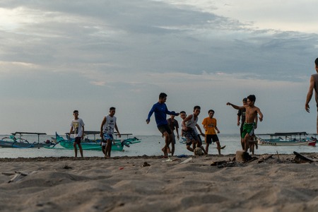 BADUNG,BALI/INDONESIA-APRIL 02 2019: Asian Teenager play football or soccer at Kuta beach with sunset or golden hours background.The sky is cloudy and some traditional boat which is called jukung, anchored thereのeditorial素材
