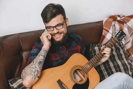 Young guitarist hipster at home holding guitar phone callの写真素材